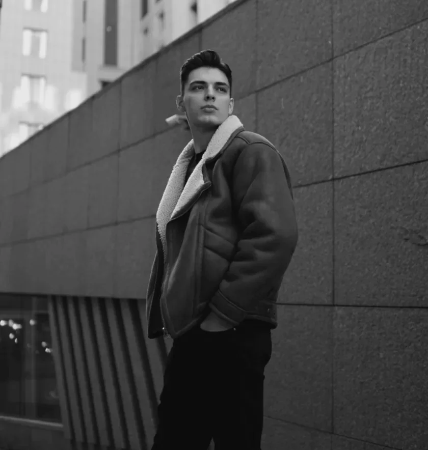 Man in a shearling-collar jacket against a stone wall, black and white